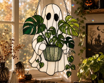 May include: A stained glass suncatcher shaped like a white ghost holding a green potted monstera plant. The ghost has two black oval eyes and is suspended by a black metal chain. The plant features multiple green leaves and trailing vines.