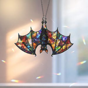 May include: A hanging ornament shaped like a bat with wings featuring a multi-colored stained glass pattern in shades of red, blue, yellow, and green. The bat is black with orange eyes and is suspended from a thin silver-toned metal chain.