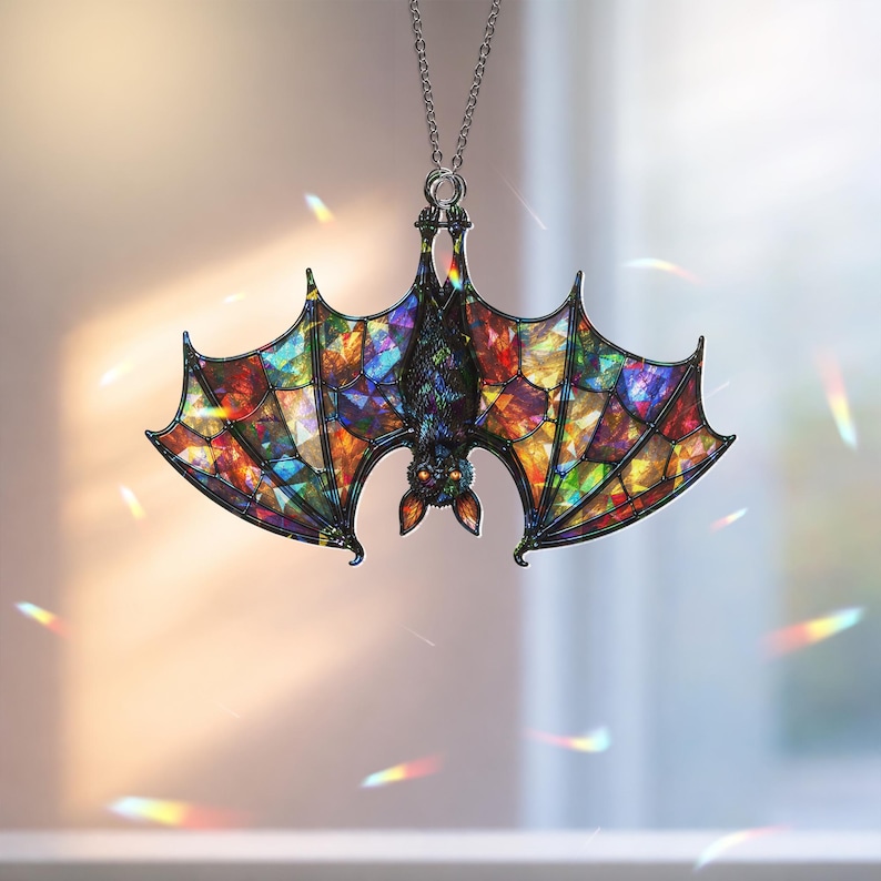 May include: A hanging ornament shaped like a bat with wings featuring a multi-colored stained glass pattern in shades of red, blue, yellow, and green. The bat is black with orange eyes and is suspended from a thin silver-toned metal chain.
