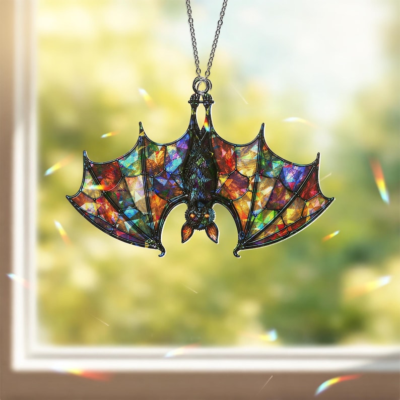 May include: A hanging ornament shaped like a bat with wings featuring a multi-colored stained glass pattern. The bat is depicted in a hanging position with its head facing downward, suspended by a thin silver-toned metal chain.