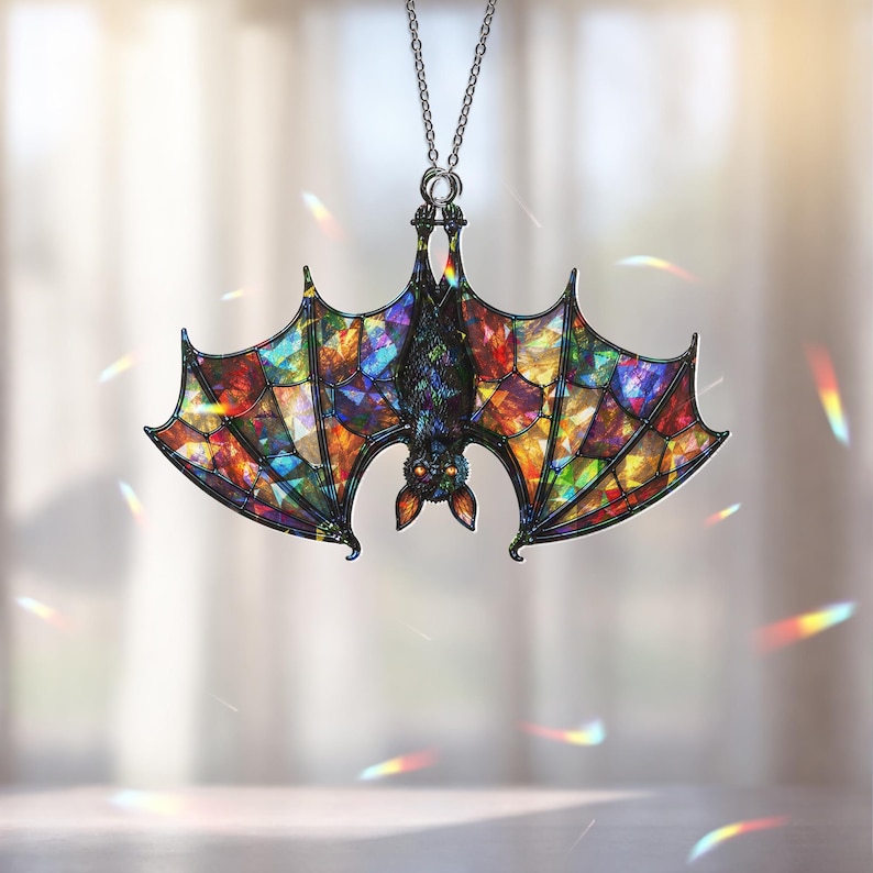 May include: A hanging ornament shaped like a bat with wings featuring a multi-colored stained glass pattern. The bat is depicted in a hanging position with dark, textured fur and glowing orange eyes, suspended from a thin silver-toned metal chain.