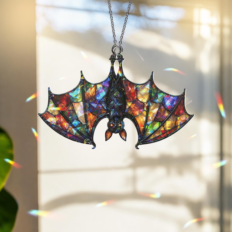 May include: A hanging ornament shaped like a bat with wings featuring a multi-colored stained glass effect in shades of red, blue, yellow, and green. The bat has a dark textured body and bright orange eyes, suspended from a silver-toned metal chain.