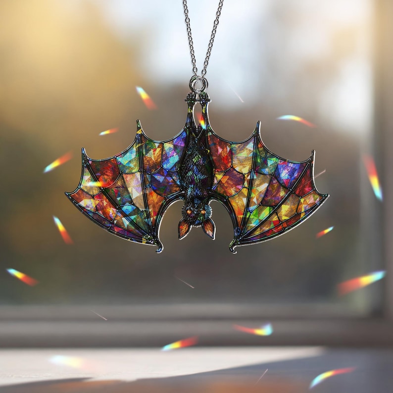 May include: A hanging ornament shaped like a bat with stained glass style wings featuring a mosaic of vibrant colors including red, blue, green, and yellow. The bat has a dark body, pointed ears, and orange eyes, suspended by a thin silver chain.