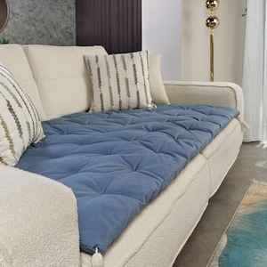 Könnte beinhalten: Ein cremefarbenes Sofa mit einem blau gesteppten Kissen und dekorativen Kissen. Das Sofa hat einen strukturierten Stoff und ein Quastendetail. Eine goldene Stehlampe steht in der Nähe.