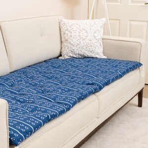 May include: A blue patterned cushion cover rests on a cream-colored sofa. The cushion cover features a repeating white geometric design. A white decorative pillow and a floor lamp are also visible.