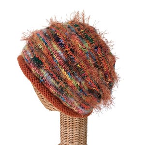 May include: A multi-colored knit beanie featuring a textured, fuzzy surface with variegated yarn in shades of orange, green, blue, and yellow. The hat has a solid, ribbed orange brim at the base, providing a distinct contrast to the variegated crown.