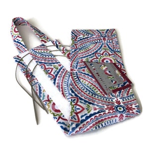 May include: A white fabric bag with a paisley pattern in shades of blue, red, and green. The bag has a handle and a pocket on the front. The pocket contains a metal needle gauge and several knitting needles.