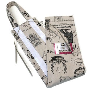 May include: Beige fabric knitting needle case with a Parisian theme. The case features a handle, white stripes, and a pocket. The fabric is printed with Eiffel Towers, stamps, and text. Knitting needles are visible inside the case.
