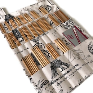 May include: A beige fabric roll-up case with black and white Eiffel Tower print and a black and white script design. The case has multiple slots for holding knitting needles. The case also includes a metal needle gauge with a red and white label.