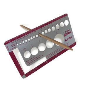 May include: A silver knitting needle gauge with a red border and a wooden knitting needle. The gauge has a ruler in inches and centimeters, and a series of holes for different needle sizes. The gauge is labeled "Susan Bates Knit-Chek".