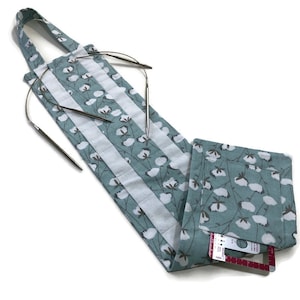 May include: A teal blue fabric needle holder with a white cotton print. The holder has a strap and a pocket for a ruler. It is filled with knitting needles.