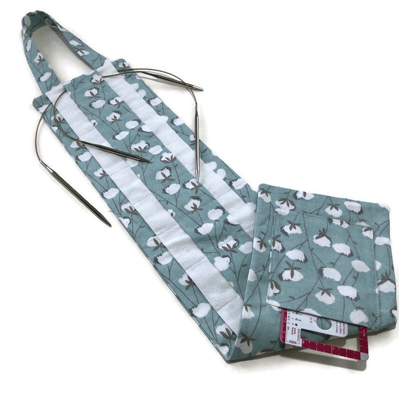 May include: A light blue fabric needle holder with white cotton flower print. The holder has a strap and a pocket. It is holding a set of circular knitting needles.