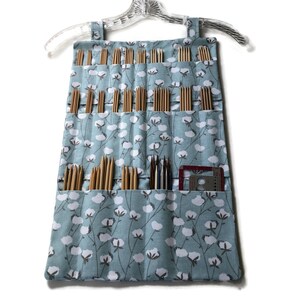 May include: A light blue fabric hanging organizer with white cotton flower print, designed to hold knitting needles. The organizer has multiple pockets to store different sizes of needles.