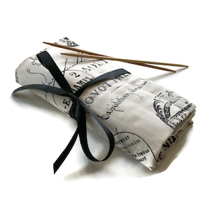 May include: A white fabric roll with black text and a black ribbon tied around it. The fabric has a vintage French design with the text "Expédition vers l'Amérique" and "2 Aout 1817". Two wooden knitting needles are inside the roll.