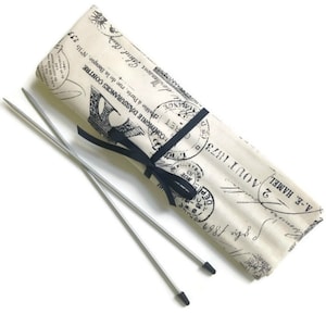 May include: A set of two silver knitting needles with black tips, wrapped in a fabric case with a black ribbon. The case has a vintage Parisian design with black and white stamps and text.