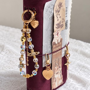 May include: A deep purple journal with a suede-like texture, adorned with a gold-toned beaded charm featuring a planet-shaped pendant. The cover includes a vertical paper insert with cat illustrations, the text "CLASSIC LEGEND," and heart-shaped charms.