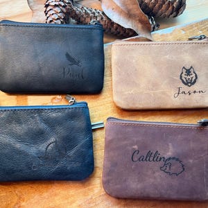 May include: Four rectangular leather zip pouches in black, tan, dark blue, and brown. Each pouch features a unique laser-engraved design: a bird with the name Paul, a wolf with the name Jason, a compass, and a hedgehog with the name Caitling.