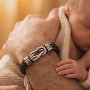 May include: A black leather bracelet with a silver infinity knot design. The bracelet is worn on a wrist, with a baby in the background. The bracelet has a braided leather band and silver accents.