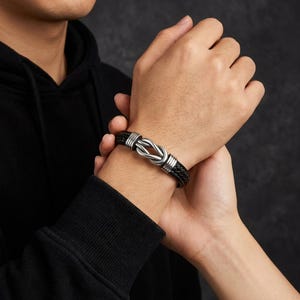 May include: Black leather bracelet with a silver knot design. The bracelet features a braided leather band and a polished silver knot centerpiece. The bracelet is secured with silver accents on each side of the knot.