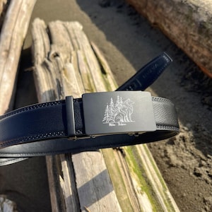May include: A black belt with a textured strap and a rectangular matte black buckle. The buckle features a laser-engraved graphic of a wolf sitting in front of pine trees with the text "Miho Robin" written in a cursive script below the image.