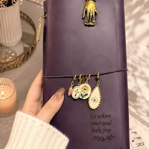 Peut inclure: Un carnet violet foncé doté d'une pince dorée en forme de main sur le dessus et de quatre breloques suspendues, dont une croix, un tournesol et un œil. La couverture porte l'inscription "Go where your soul feels free" et un motif de feuilles.
