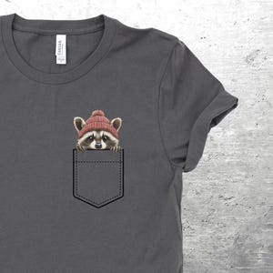 May include: A dark gray t-shirt with a front pocket featuring a raccoon wearing a red beanie hat. The raccoon is peeking out of the pocket.