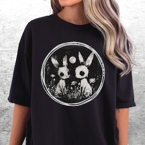 Op de afbeelding: Zwart oversized T-shirt met een grafische print van twee witte konijnen met zwarte ogen in een cirkel. De konijnen zijn omringd door witte bloemen en een halve maan.