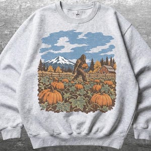Peut inclure: Un sweat-shirt gris clair à col rond avec une impression graphique représentant une créature Bigfoot brune marchant dans un champ de citrouilles. La scène inclut des citrouilles orange, une clôture en bois, une grange rouge, des sapins et une montagne enneigée.