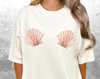 May include: A white t-shirt with a pink seashell graphic on the front. The seashells are stylized and have a watercolor effect.