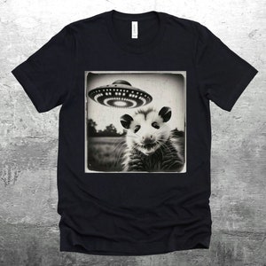May include: Black and white graphic t-shirt featuring a photo of a possum looking up at a UFO. The photo is framed in a vintage style.