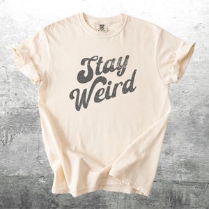 K&ouml;nnte beinhalten: Cremefarbenes T-Shirt mit dem Aufdruck "Stay Weird" in verblichenen, dunkelgrauen Buchstaben. Das Shirt hat kurze &Auml;rmel und einen Rundhalsausschnitt und ist aus einem weichen, bequemen Material gefertigt.