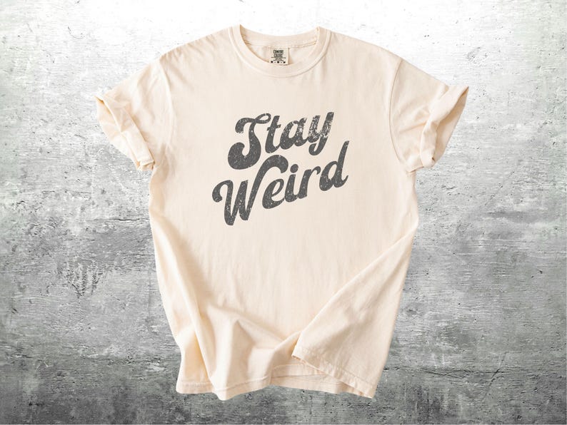 K&ouml;nnte beinhalten: Cremefarbenes T-Shirt mit dem Aufdruck "Stay Weird" in verblichenen, dunkelgrauen Buchstaben. Das Shirt hat kurze &Auml;rmel und einen Rundhalsausschnitt und ist aus einem weichen, bequemen Material gefertigt.