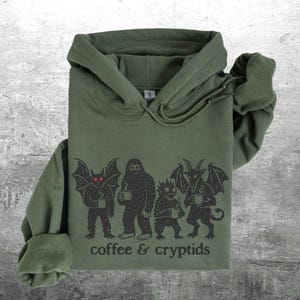 May include: Olive green hoodie with a graphic of cryptids holding mugs, with the text "coffee & cryptids" printed below. The graphic features a bat-like creature, a Bigfoot, a small monster, and a horned demon. The hoodie has a drawstring hood.
