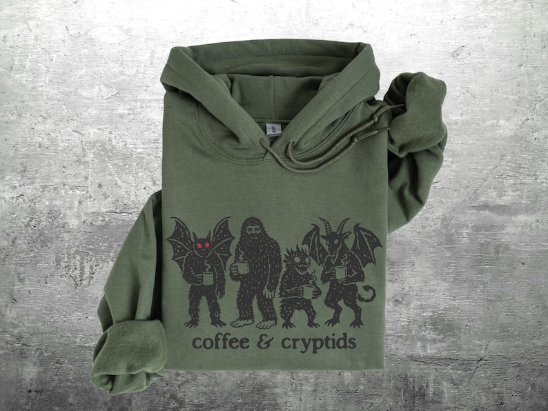 May include: Olive green hoodie with a graphic of cryptids holding mugs, with the text "coffee & cryptids" printed below. The graphic features a bat-like creature, a Bigfoot, a small monster, and a horned demon. The hoodie has a drawstring hood.