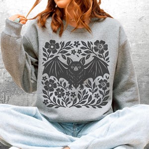 May include: A gray sweatshirt featuring a dark gray bat design surrounded by flowers and leaves. The bat has its wings spread, and the floral design is symmetrical. The sweatshirt is a casual style.