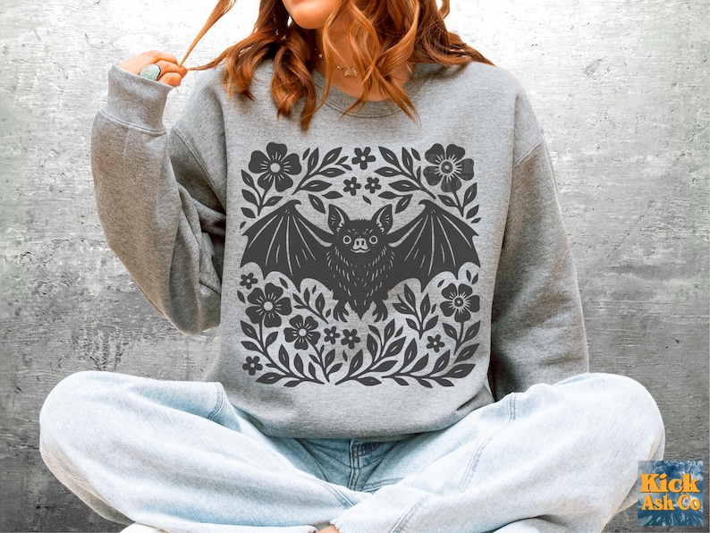 May include: A gray sweatshirt featuring a dark gray bat design surrounded by flowers and leaves. The bat has its wings spread, and the floral design is symmetrical. The sweatshirt is a casual style.