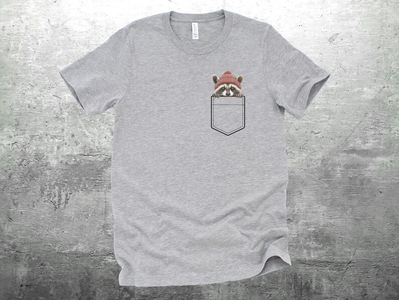 May include: A heather gray t-shirt with a pocket featuring a raccoon wearing a red beanie hat.