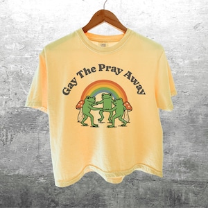May include: Yellow t-shirt with the text "Gay The Pray Away" above a graphic of three green frogs dancing under a rainbow. The frogs are positioned between two mushrooms. The shirt is hanging on a wooden hanger.
