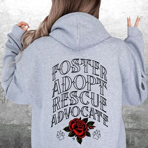 Pu&ograve; includere: Felpa con cappuccio grigia con il testo "FOSTER ADOPT RESCUE ADVOCATE" in lettere nere. Sotto il testo c'&egrave; una rosa rossa con foglie verdi e due impronte di zampa.