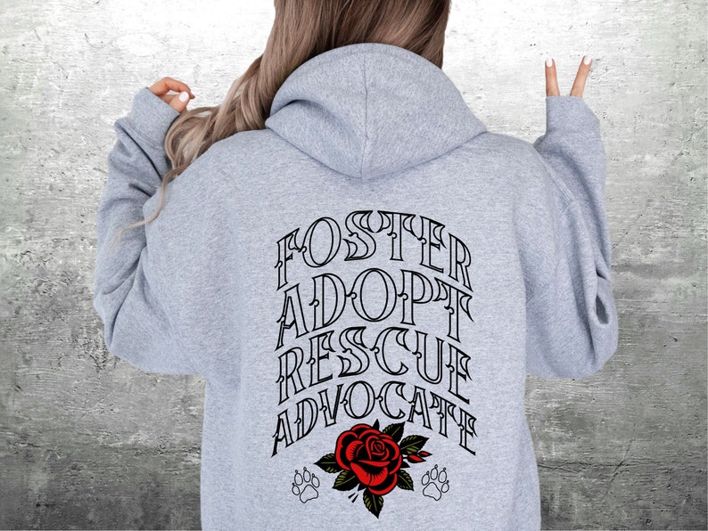 Pu&ograve; includere: Felpa con cappuccio grigia con il testo "FOSTER ADOPT RESCUE ADVOCATE" in lettere nere. Sotto il testo c'&egrave; una rosa rossa con foglie verdi e due impronte di zampa.