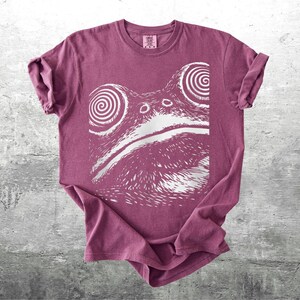 May include: A maroon t-shirt featuring a white graphic of a frog's face with spiral eyes. The shirt has rolled-up sleeves and a crew neck. The frog design is centered on the front of the shirt.