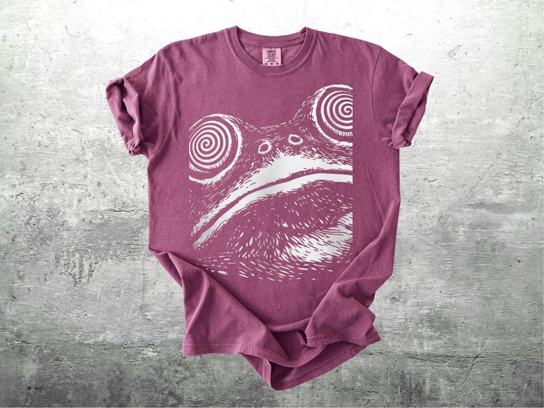 May include: A maroon t-shirt featuring a white graphic of a frog's face with spiral eyes. The shirt has rolled-up sleeves and a crew neck. The frog design is centered on the front of the shirt.