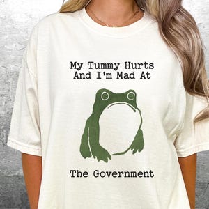 May include: White t-shirt with a green frog graphic and the text "My Tummy Hurts And I'm Mad At The Government".