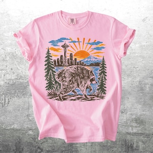 May include: A light pink t-shirt featuring a graphic print of a raccoon walking in front of a landscape. The scene includes the Seattle Space Needle, a city skyline, a mountain, pine trees, and a sunburst design in shades of blue, orange, and black.