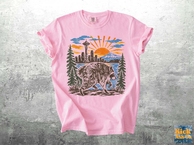May include: A light pink t-shirt featuring a graphic print of a raccoon walking in front of a landscape. The scene includes the Seattle Space Needle, a city skyline, a mountain, pine trees, and a sunburst design in shades of blue, orange, and black.