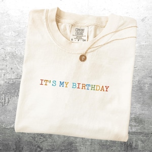 Puede incluir: Una camiseta blanca con el texto "IT'S MY BIRTHDAY" en colores arco&iacute;ris. La camiseta tiene una peque&ntilde;a etiqueta que dice "COMFORT COLORS" y "S - P - CN".