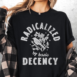 May include: Black t-shirt with the words "RADICALIZED" above a bird and floral design, and "by basic DECENCY" below. The shirt is worn with a black and tan plaid flannel.