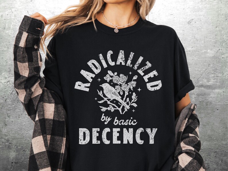 May include: Black t-shirt with the words "RADICALIZED" above a bird and floral design, and "by basic DECENCY" below. The shirt is worn with a black and tan plaid flannel.