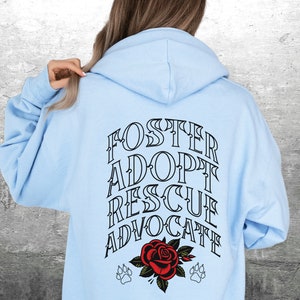 Pu&ograve; includere: Felpa con cappuccio blu chiaro con le parole "FOSTER ADOPT RESCUE ADVOCATE" in nero sul retro. Un rosa rossa con due impronte di zampa si trova sotto il testo.