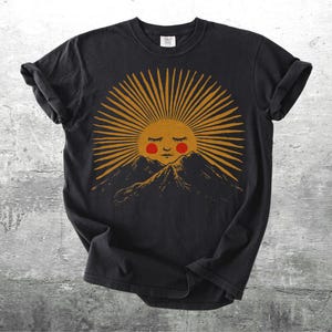 K&ouml;nnte beinhalten: Schwarzes T-Shirt mit einer Grafik einer l&auml;chelnden Sonne mit geschlossenen Augen und rosigen Wangen, die goldene Strahlen &uuml;ber einer Bergkette ausstrahlt. Das Design hat eine Vintage-&Auml;sthetik, handgezeichnet, f&uuml;r den l&auml;ssigen Gebrauch.